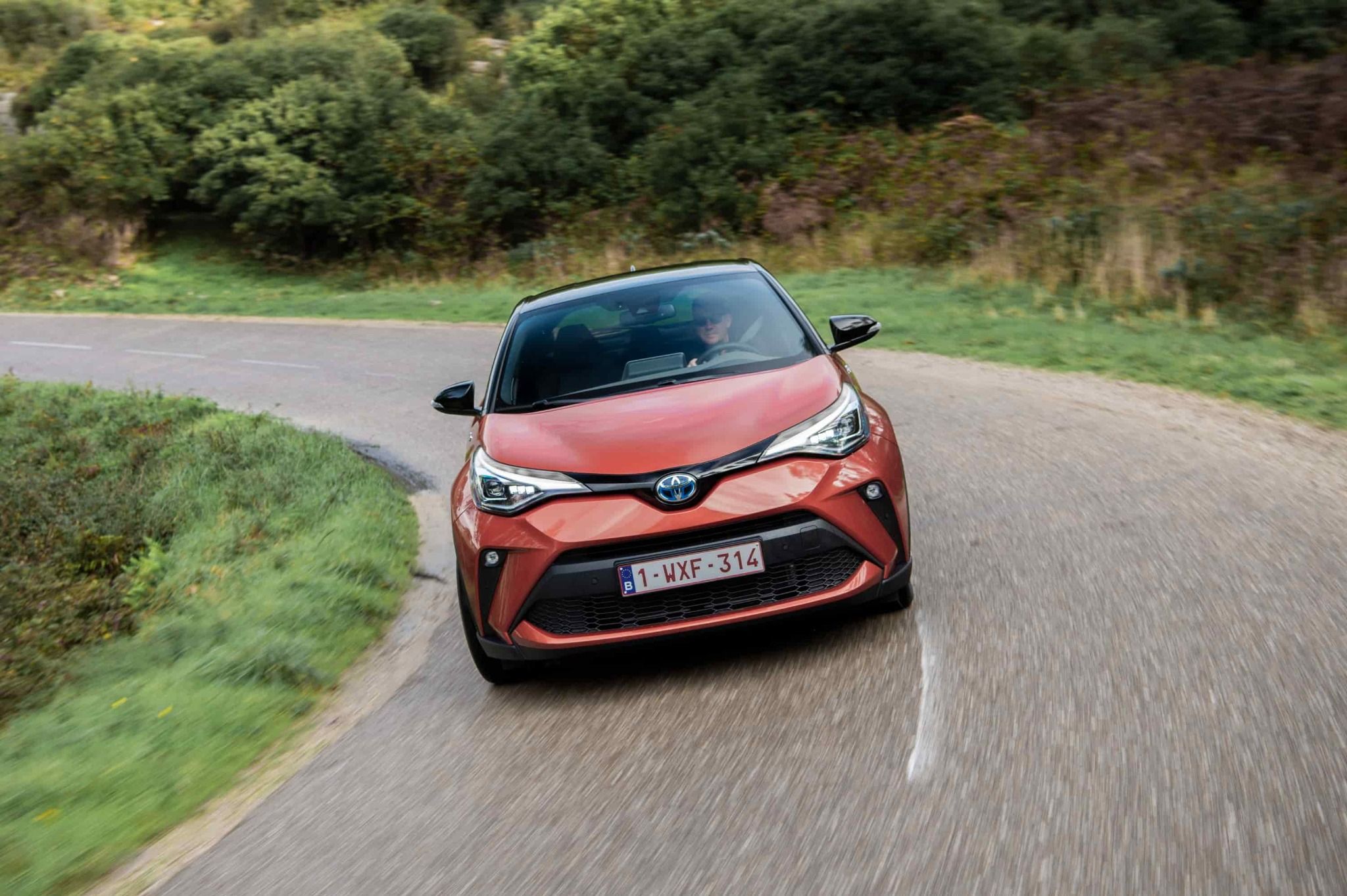 orange toyota c hr driving on a country road with trees in the background