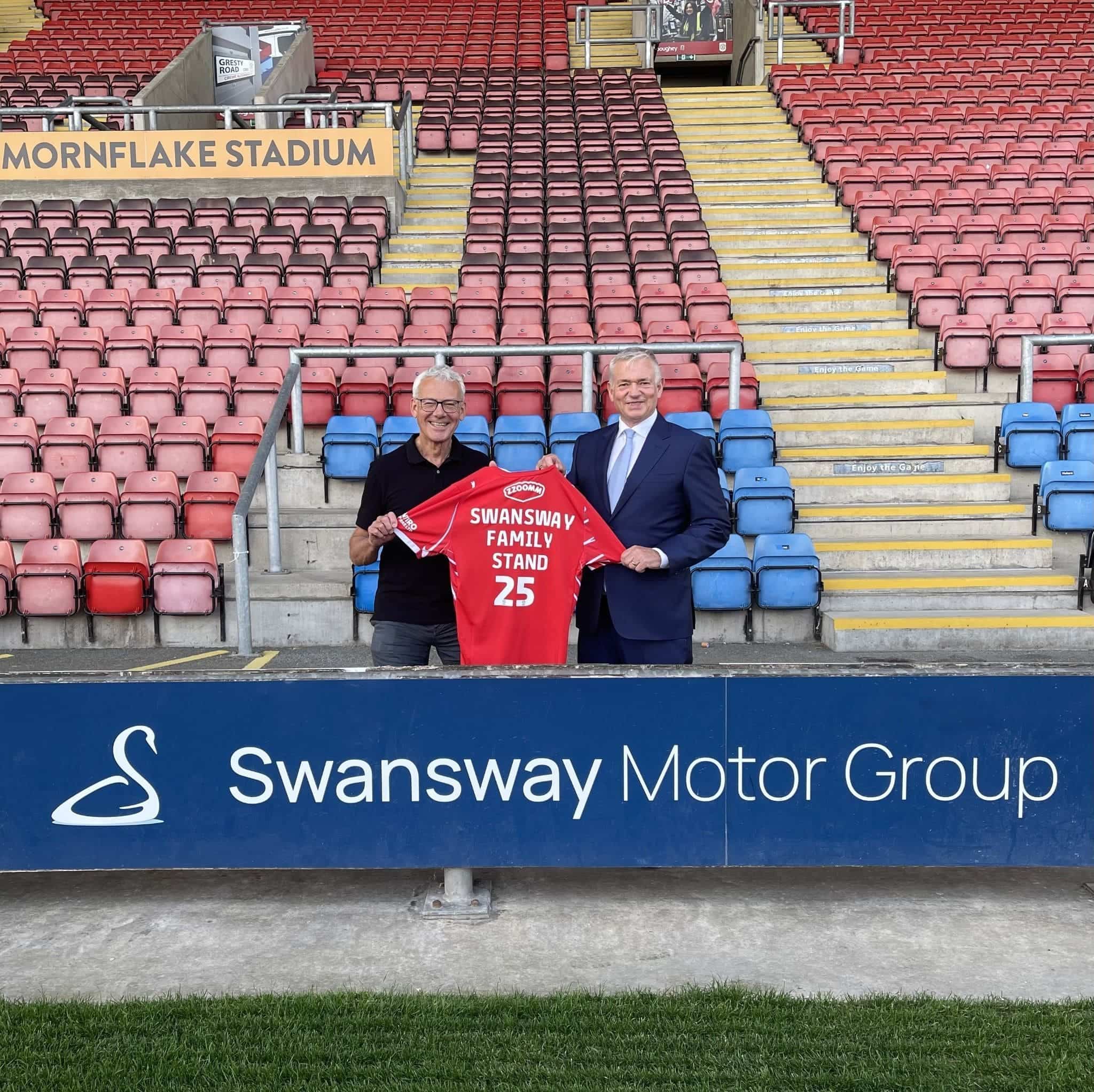 two men holding a football shirt in front of a swansway sign in a football stand