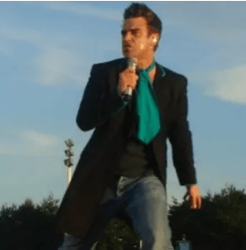 Robbie Williams singing into a microphone with a blackk coat, jeans, black t-shirt and big blue tie